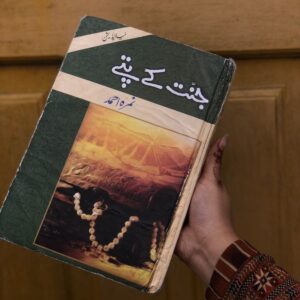 Hand holding an Urdu Islamic novel ‘Jannat Ke Pattay’ by Nemrah Ahmed with green cover, tasbeeh image, and wooden background.”