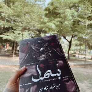 “Hand holding the Urdu novel ‘Bahl’ by Mehar-un-Nisa Shah Meer in an outdoor park with trees in the background, dark themed book cover with bird and crown artwork.”